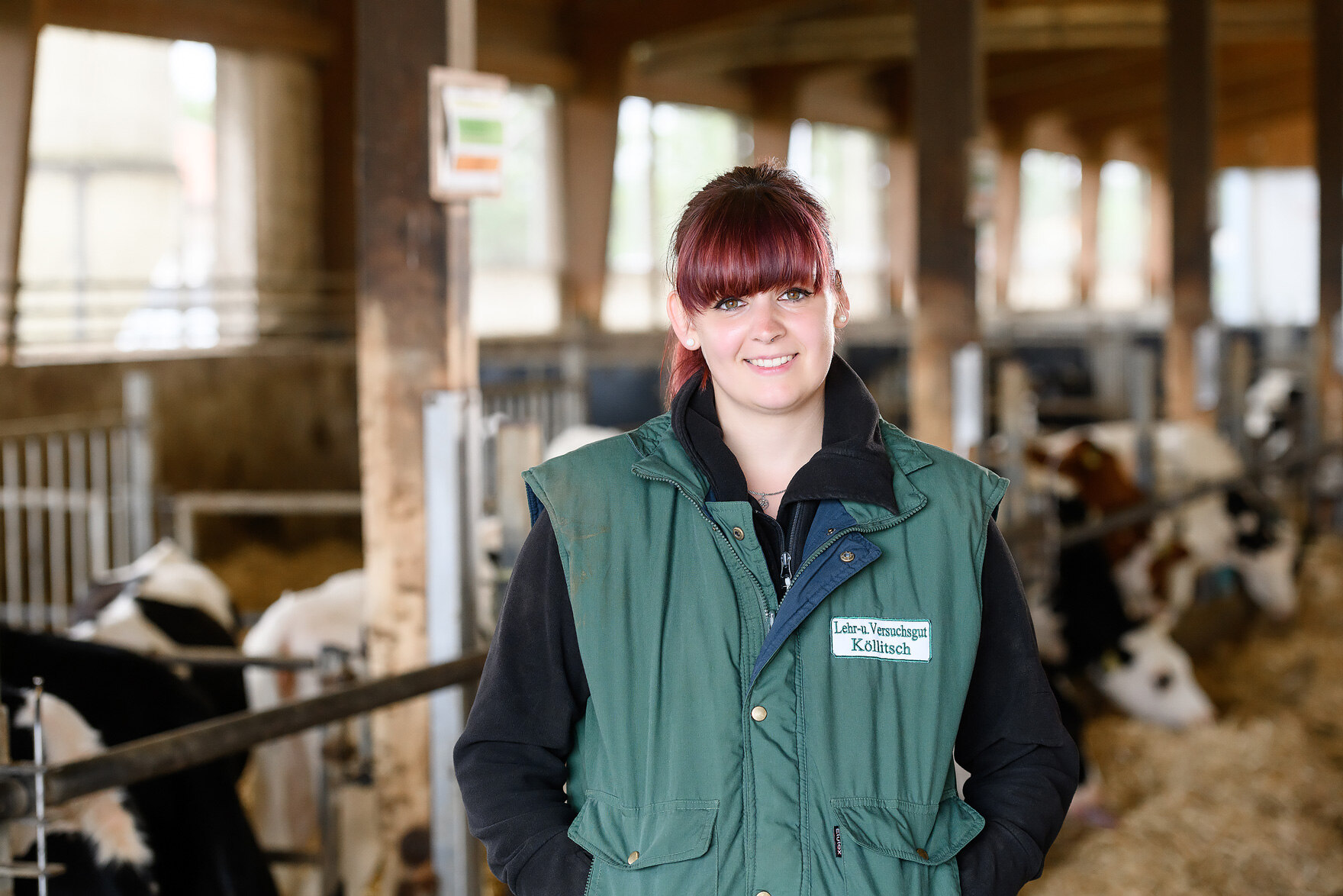 Landwirtin in Arbeitskleidung steht im Rinderstall und lächelt in die Kamera. 