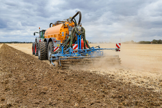 Traktor fährt mit Güllewagen über das Feld. Gülle wird in den Boden eingearbeitet. 