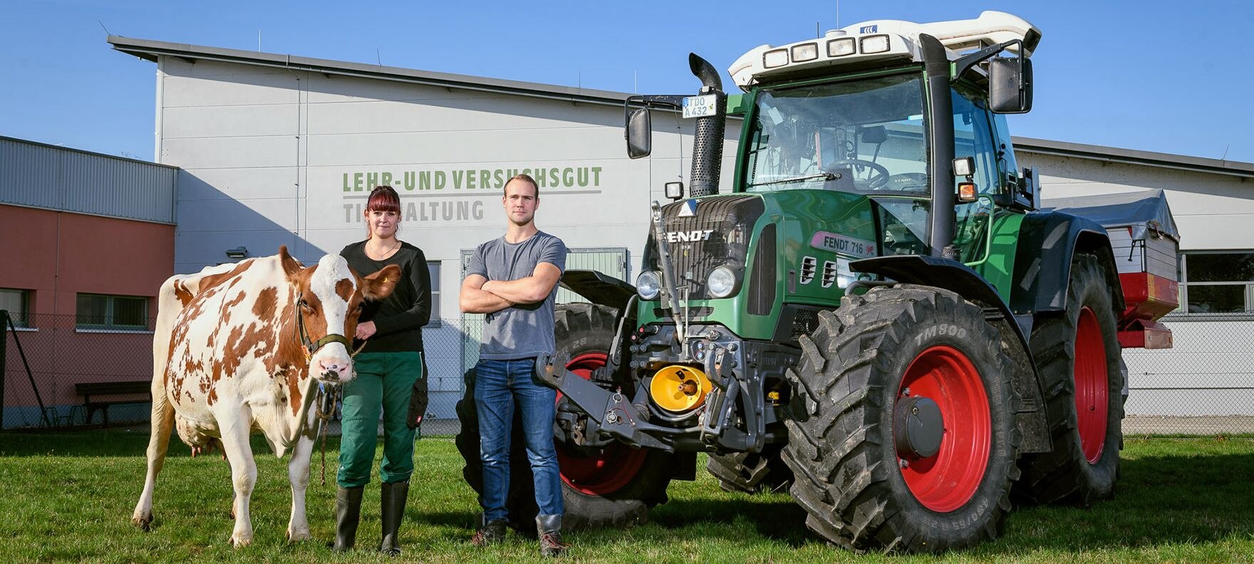 Eine Landwirtin und ein Landwirt stehen mit einer Kuh und neben einem Traktor vor einem Gebäude des LVG Köllitsch.